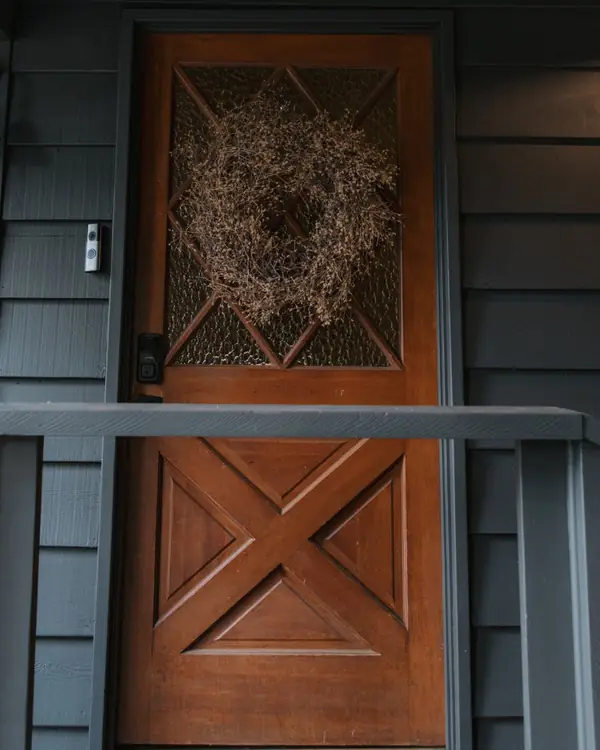 A wooden front door with a dried wreath, on a dark porch